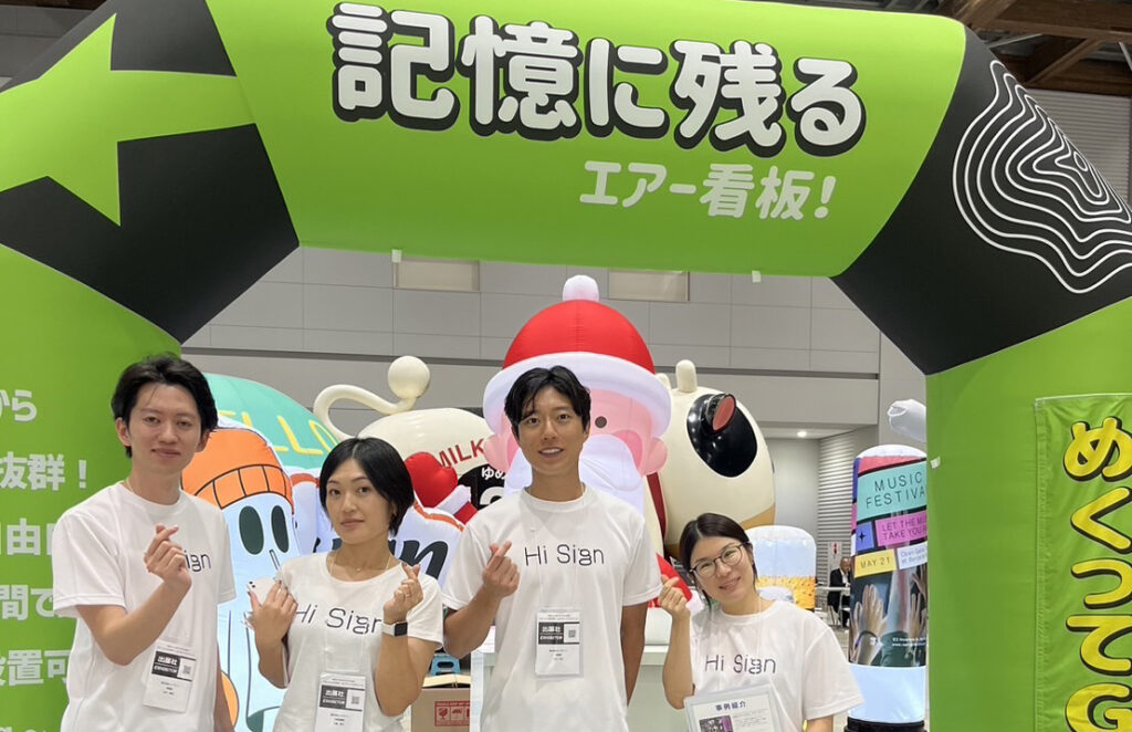 エアー看板をバックにポーズを取る4人のスタッフ